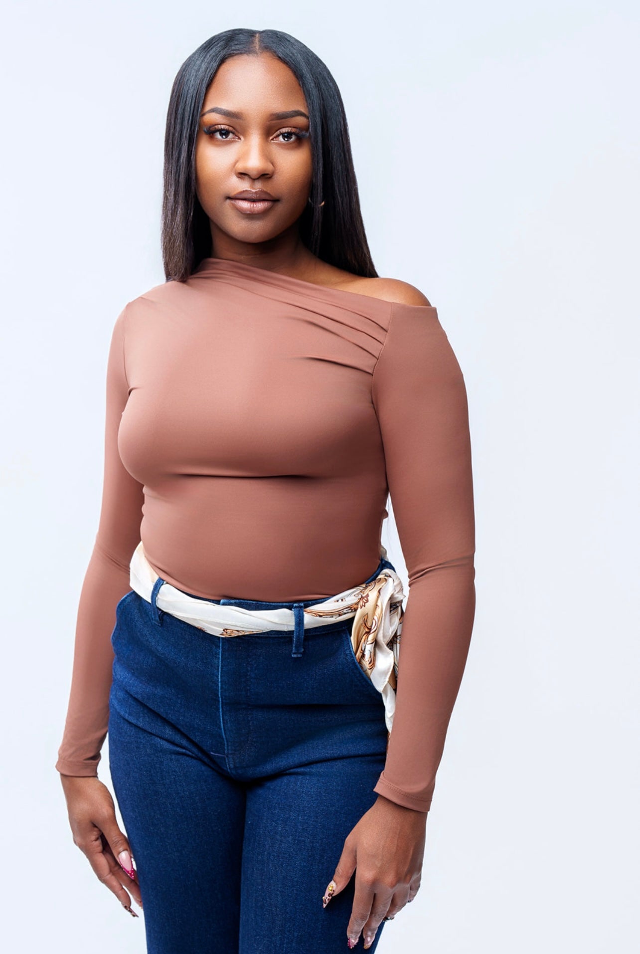 Woman wearing a brown off-shoulder top and blue jeans against a light gray background