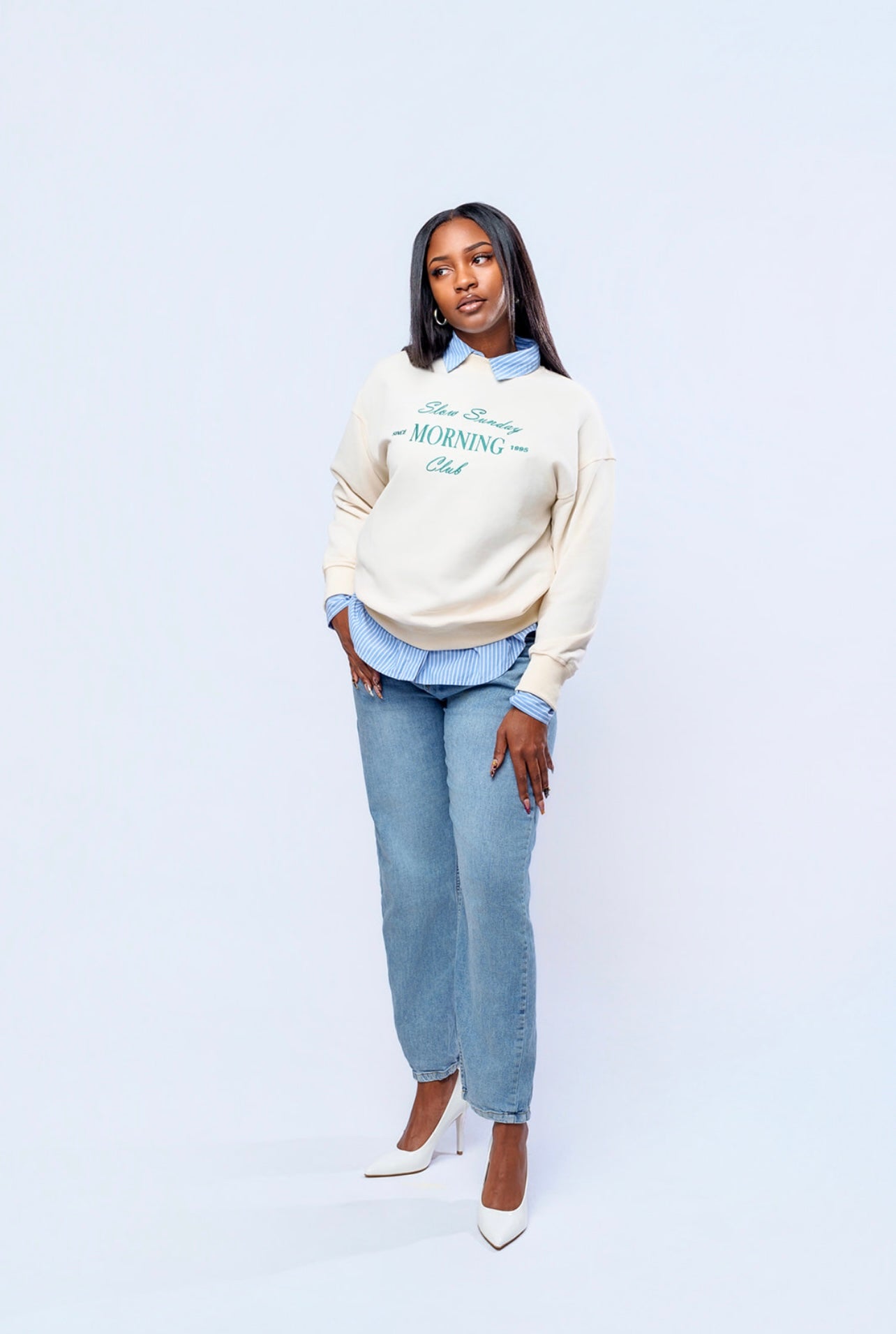 Woman wearing a beige sweater with text, light blue shirt, and jeans on a white background
