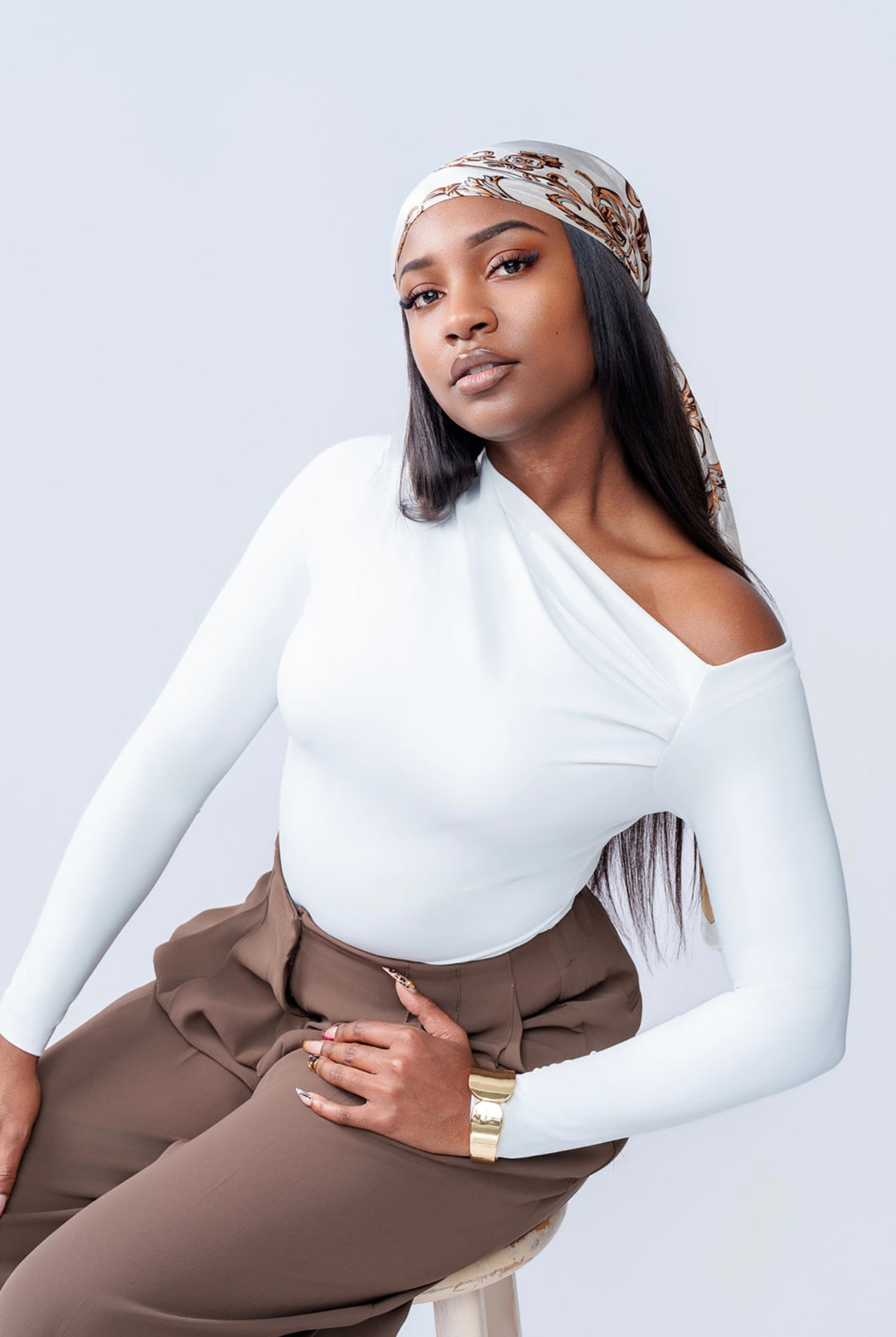 Woman wearing a white long-sleeve top and brown pants with a patterned headscarf on a light gray background
