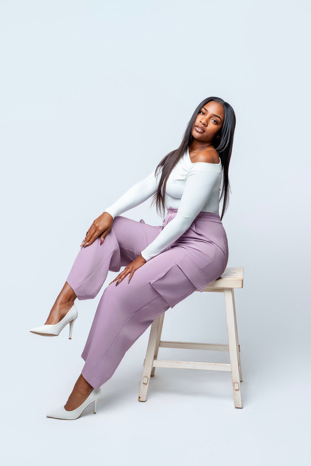 Woman in a white top and lavender pants sitting on a stool against a light gray background