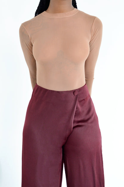 Person wearing a fitted, sheer nude long-sleeve top tucked into high-waisted, wide-leg maroon satin pants, standing against a plain white background.