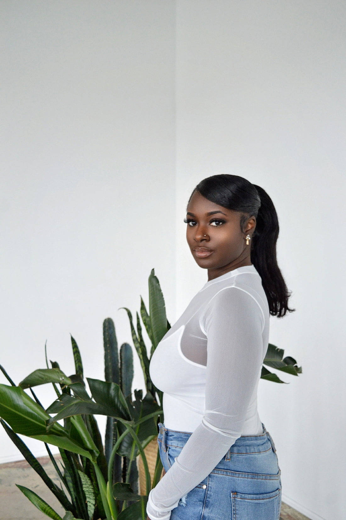 Person wearing a fitted, sheer white long-sleeve top tucked into light wash distress denim jeans standing against a plain white background.