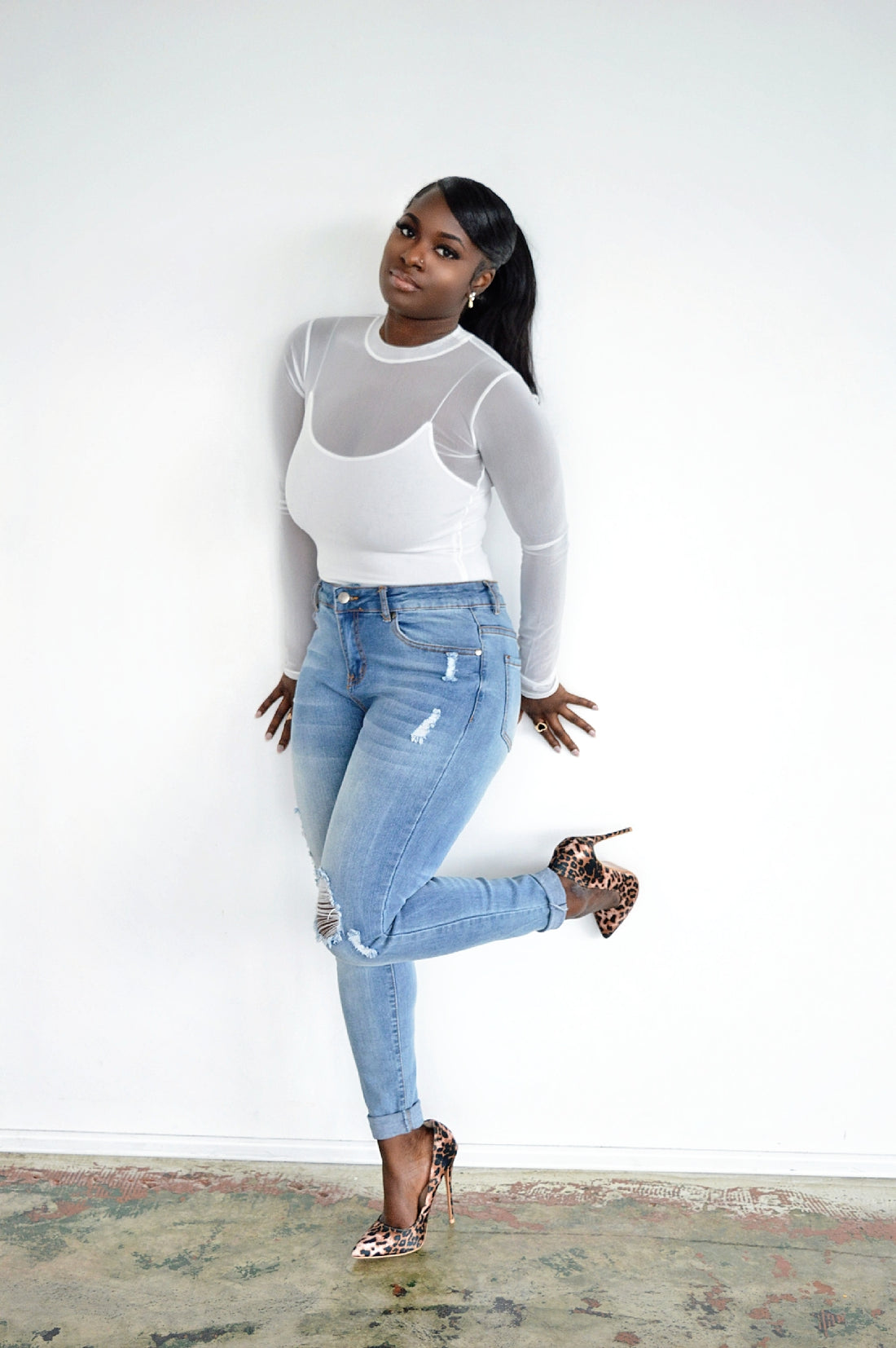 Person wearing a fitted, sheer white long-sleeve top tucked into light wash distress denim jeans standing against a plain white background wearing.